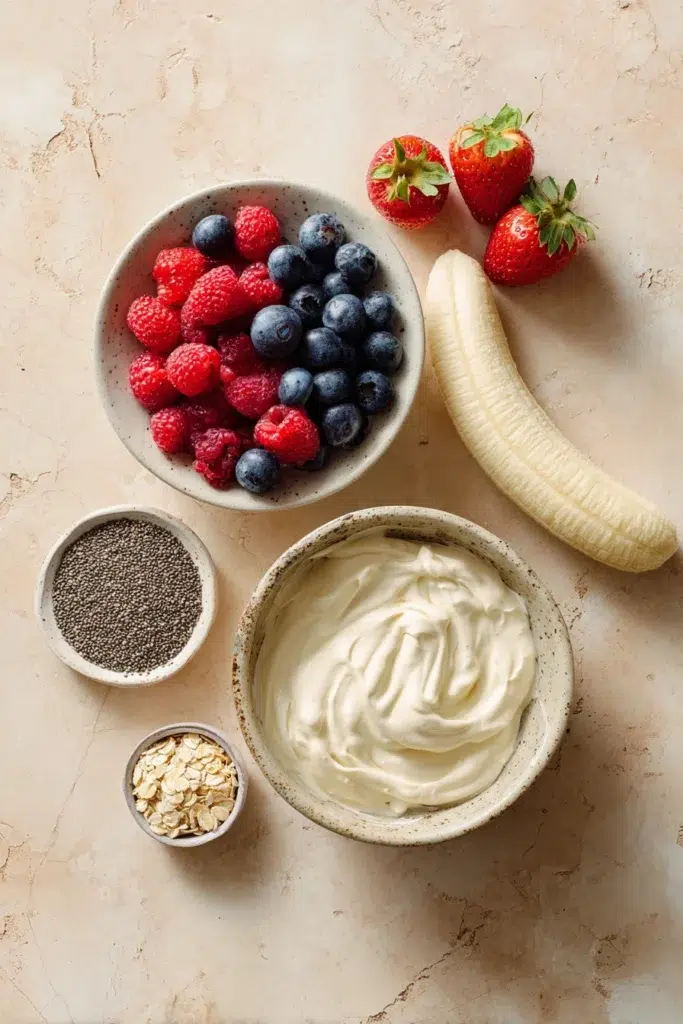 frozen yogurt bark for weight loss. 2 Flat lay of frozen yogurt bark ingredients: Greek yogurt, banana, fresh strawberries, raspberries, blueberries, chia seeds, and oats on a marble background"